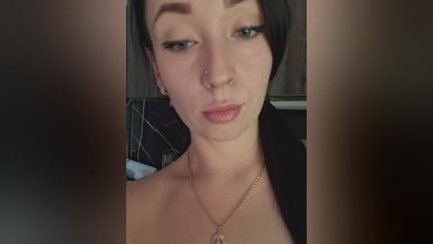 A close-up video of a young woman with short dark hair, light skin, and a small nose piercing. She wears a gold chain necklace and a black top. The background is blurred, suggesting an indoor setting.