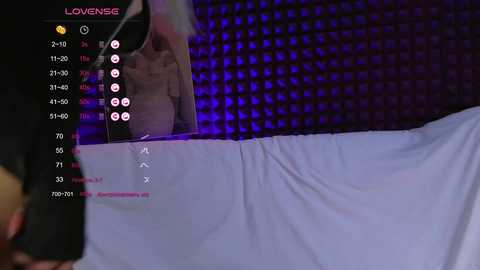 A video shows a live stream with a blurred person in a brown jacket, speaking into a microphone against a grid background. The lower part displays a bed with white sheets and a man's arm.