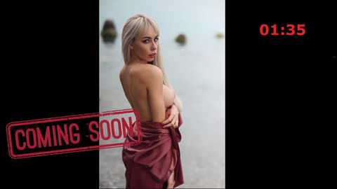 Video of a topless blonde woman with fair skin, wearing a maroon robe, standing seductively against a blurred water background. Text reads \"COMING SOON\" in bold red letters.