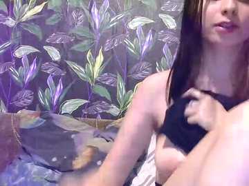 Media: Video of a young woman with long brown hair, wearing a black top, sitting in a room with a purple floral wallpaper. Her expression is neutral.