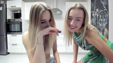 Media: Video of two young blonde women in a modern kitchen. One topless, one in a green cheerleader outfit, both smiling. White cabinets, black fridge, chalkboard wall, stainless steel oven.