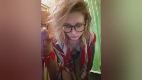 A blurry video of a young woman with light skin and blonde hair, wearing glasses and a striped shirt, leaning against a wall in a dimly lit room.
