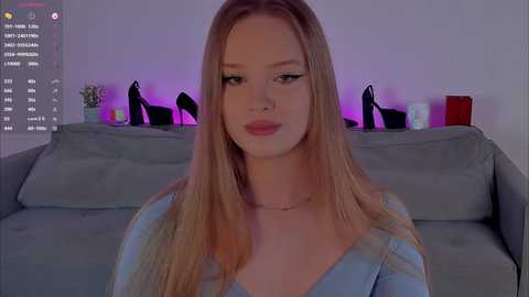 Video of a young Caucasian woman with long blonde hair, wearing a light blue top, sitting on a gray couch with black high heels on a shelf behind her.