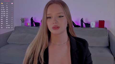 A video of a young, fair-skinned woman with long blonde hair and a black blazer, sitting on a gray couch in a dimly lit room with purple ambient lighting.