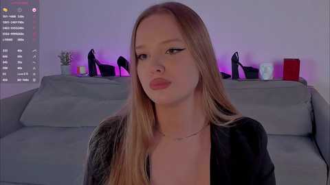 A video of a young, fair-skinned, blonde woman with long hair, wearing a black blouse and necklace, sitting on a gray couch. The background features a purple-lit room with black high heels and potted plants.