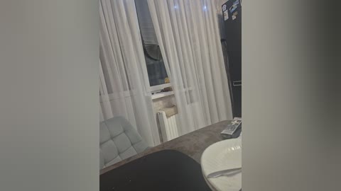 Video of a modern, minimalistic bathroom with a white sink, gray countertop, white toilet, light blue upholstered chair, white curtains, and a mirror. The room has a clean, uncluttered appearance.