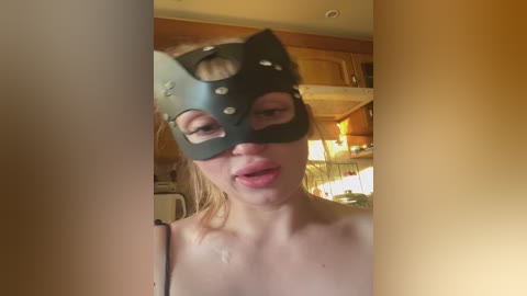 Video of a fair-skinned woman in a black leather cat mask with holes, wearing a black top, indoors.