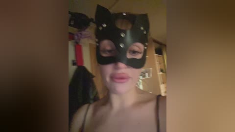 A video of a fair-skinned woman wearing a black cat mask with studs and a spaghetti-strap top, standing in a dimly lit room with beige walls and a shelf visible in the background.