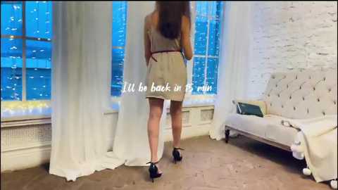 Video of a woman in a beige dress standing with her back to the camera, looking out large windows. Text reads, \"I'll be back in 5 min.\