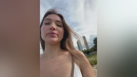 Media: Video of a topless woman with long brown hair, wearing a white dress, standing outdoors with a cloudy sky and tall buildings in the background.