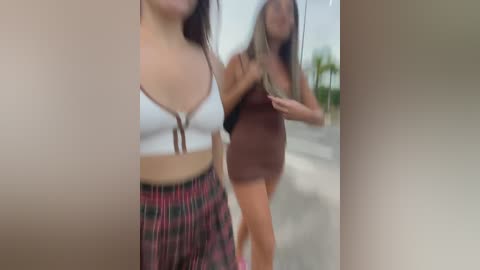 Media: Video of two women walking outdoors. The woman in the foreground wears a white crop top and plaid shorts. The blurred background shows a second woman in a brown dress.