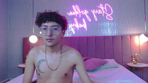 Video of a young, shirtless, slim, light-skinned man with curly hair, wearing glasses, and a necklace, seated on a bed with pink pillows, illuminated by neon \"Follow your dreams\" sign and bedside lamps.