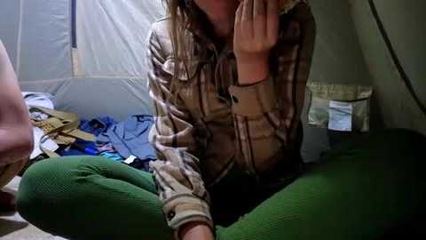 Video of a person with shoulder-length blonde hair, wearing a plaid shirt and green corduroy pants, sitting cross-legged inside a tent, with scattered camping gear and a sleeping bag in the background.