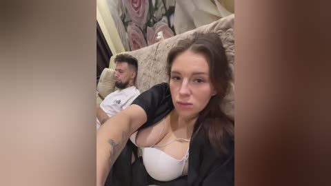 Media: Video of a woman with long brown hair and a tattooed arm, wearing a white bra and black top, sitting next to a man in a white shirt on a beige couch in a room with floral wallpaper.