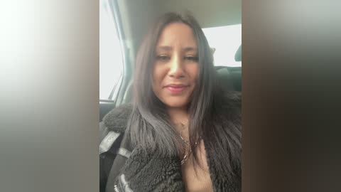Video of a Latina woman with long, straight, dark hair, wearing a gray fur coat and seated in a car, smiling, with a blurred background.