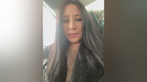 Video of a young woman with long, straight dark hair and a medium complexion, wearing a black textured jacket and a necklace. She sits in a car, with a bright light source creating a soft glow in the background.