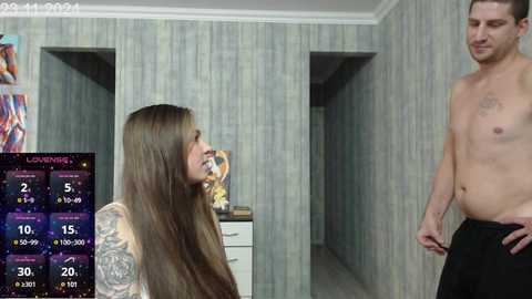 Video of a topless, muscular man in black pants, standing in a hallway with wallpaper. A tattooed woman with long hair kneels, holding a drink, in front of a digital clock displaying 30\u00b0C.