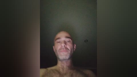 Video of a bald, middle-aged Caucasian man with a light beard, standing shirtless against a dark, dimly lit wall. His expression is neutral.