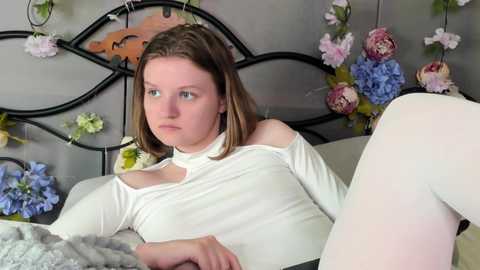 Video of a fair-skinned, young woman with shoulder-length brown hair, wearing a white off-shoulder top and white tights, sitting on a bed with floral decorations, looking thoughtful.
