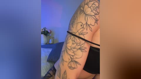 Video of a light-skinned woman with a large floral tattoo on her right arm. She wears black lingerie, standing against a blue-lit wall with a vase of flowers in the background.