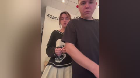 Media: A video shows a young woman in a black sweater with a white skull design and a white skirt, standing in a hallway, with a man in a black shirt in the foreground.