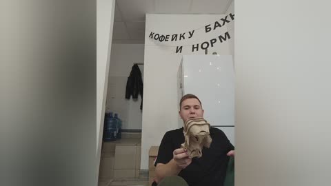 Media: A video captures a man in a black t-shirt holding a beige toy dinosaur in a minimalist room with white walls, a white refrigerator, and a blue backpack.