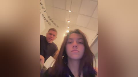 Media: Video of a young woman with long brown hair and a light complexion, wearing a dark jacket, looking at the camera with a neutral expression. Behind her, a man with short hair and a light complexion is visible, also looking at the camera.