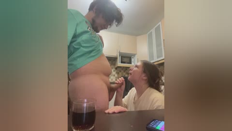 Video: A man with a beer belly, wearing a green shirt, stands in a kitchen. A woman, seated, gazes up at him. The background shows a microwave, cabinets, and a dark countertop with a glass of beer.