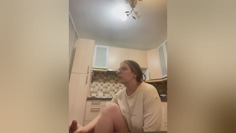 A video of a woman with glasses, light skin, and brown hair in a beige robe, sitting on a kitchen floor with beige cabinets, a tiled backsplash, and a ceiling light.