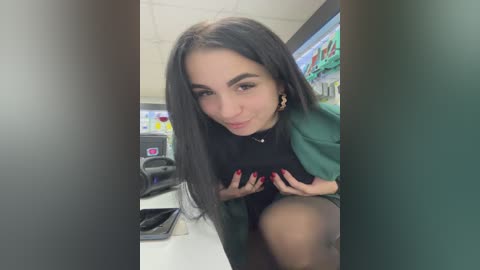 Video of a young woman with long, straight black hair, wearing a green blazer over a black top, sitting at a desk, smiling, with a phone in her hands.