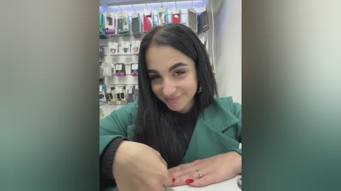 Video of a smiling Latina woman with long black hair, wearing a green lab coat, seated in a pharmacy, surrounded by shelves of products.