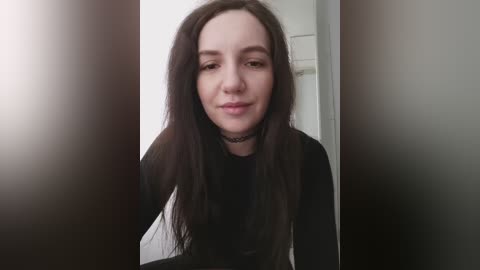 Video of a young woman with long, straight dark hair and pale skin, wearing a black choker and long-sleeved top. She has a neutral expression. The background is blurred, featuring a white wall.