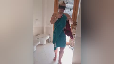 Video of a person, wearing a teal towel, taking a mirror selfie in a bathroom with white walls, a toilet, and a wooden door with blinds.
