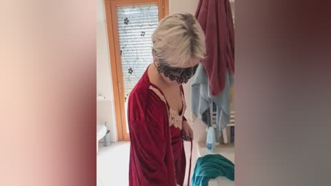 Video of a short-haired, light-skinned woman in a red robe with a lace collar, wearing a black mask, washing a blue and white object in a bathroom with beige walls and wooden-framed windows.