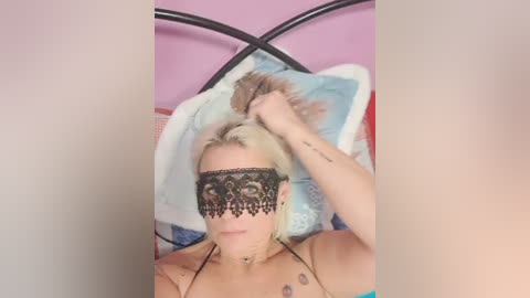 Video of a blonde woman with a black lace mask, holding her hair, lying on a bed with a black metal headboard. She has tattoos on her arms and chest.