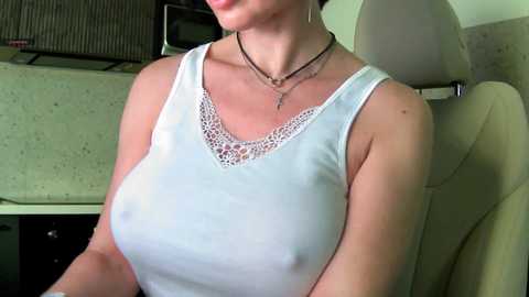 Media: Video of a light-skinned woman with a curvy figure wearing a sheer white lace top that reveals her large breasts and nipples, seated in a beige car. She wears a black necklace.