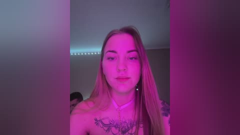 Media: Video of a young Caucasian woman with long blonde hair, wearing a necklace, standing in a dimly lit room with purple lighting. She has tattoos on her chest and shoulders.