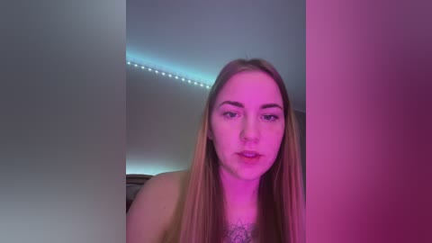 Media: Video of a young Caucasian woman with long, straight, light brown hair, wearing a light-colored top, standing against a backdrop of a dimly lit room with purple and blue lighting.