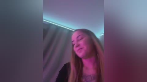 Media: A video of a young woman with long, light brown hair, wearing a black top, smiling softly. The background features a dimly lit, curtain-covered room with soft, purple lighting.