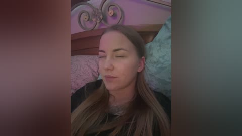 Media: Video of a fair-skinned, blonde-haired woman with closed eyes, wearing a black shirt, lying on a bed with a floral-patterned pillow and wooden headboard, in a dimly lit room.