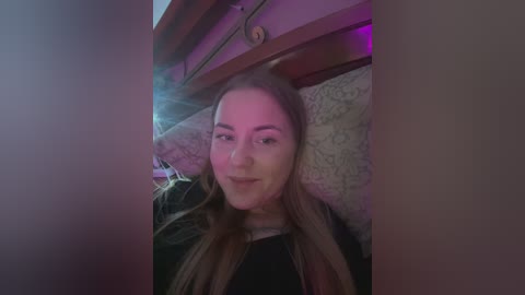 Media: A video of a young Caucasian woman with long brown hair, wearing a black top, smiling slightly, sitting in a dimly lit room with purple and white lighting, and wooden beams in the background.