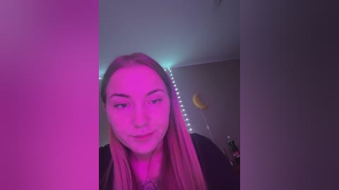Media: Video of a young woman with long, straight blonde hair, wearing a black top, standing in a dimly lit room with pink and purple LED lights. The background features a yellow moon-shaped lamp and a small potted plant.