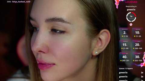 Media: Video of a close-up of a young woman with light skin, straight brown hair, and a light pink lipstick. The background shows a dimly lit room with a green screen overlay.