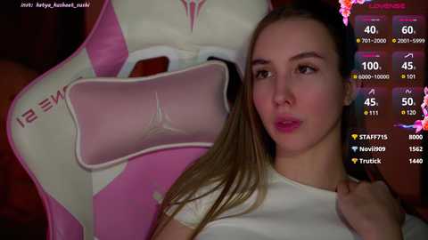 Media: Video of a young woman with fair skin and brown hair, wearing a white top, standing beside a pink and white gaming chair.
