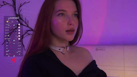 Video of a young woman with long red hair, wearing a black off-shoulder top and a choker necklace. Purple lighting highlights her fair skin, set against a minimalist background.
