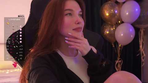 Video of a fair-skinned, long-haired woman with a serene expression, wearing a black jacket over a white top, seated in a dimly lit room with gold balloons and a calendar.