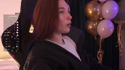 Video of a young woman with fair skin and long red hair, wearing a white top and black hoodie, sitting in a gaming chair. Background includes a black curtain, gold balloons, and a digital clock.