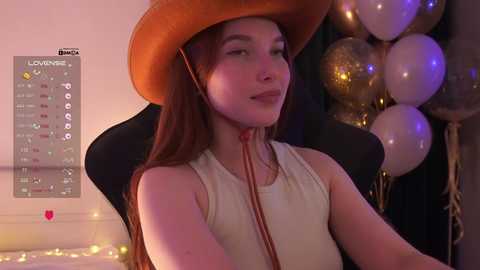Video of a smiling, fair-skinned woman with long brown hair wearing a tan cowboy hat and a white sleeveless top, seated in a black gaming chair. Background features gold and white balloons, a calendar, and soft lighting.