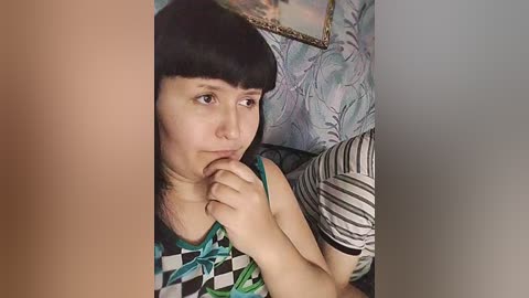 A video of a woman with straight black hair and bangs, wearing a green and black checkered top, sitting pensively on a patterned couch with a framed painting in the background.