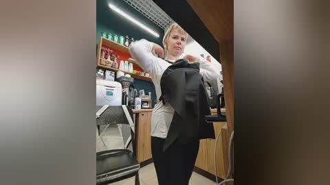Video of a blonde woman in a white shirt, black vest, and black pants, removing a black jacket in a modern, brightly-lit salon. Shelves with products and a white coffee machine are visible in the background.